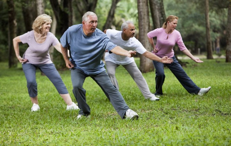 Multi-ethnic group of adults practicing tai chi in park.  Main focus on senior man (60s) in blue shirt.